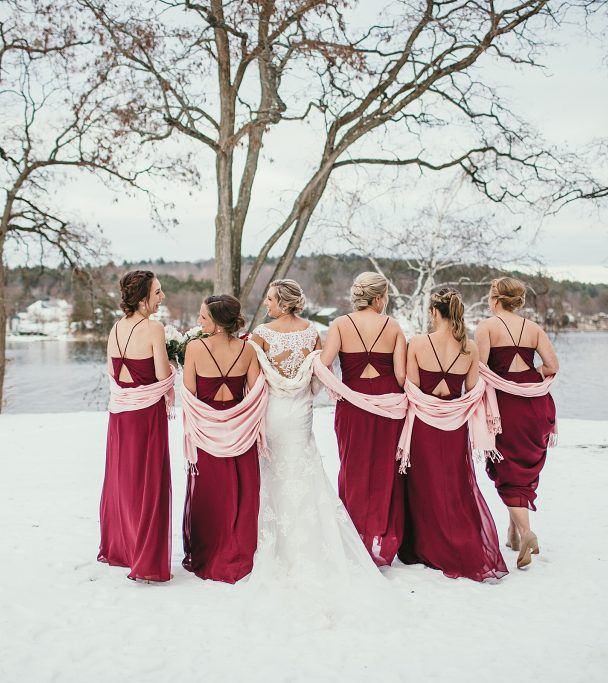 Six brides wearing matching magenta gowns stand in a snowy park, backs to camera, arms around each other, trees and a lake in the background.