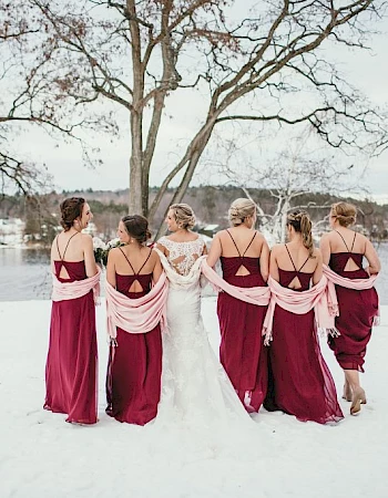 A bridal party in burgundy dresses gathers in the snowy outdoors, backs turned, tying a light wrap around shoulders near a frozen lake.