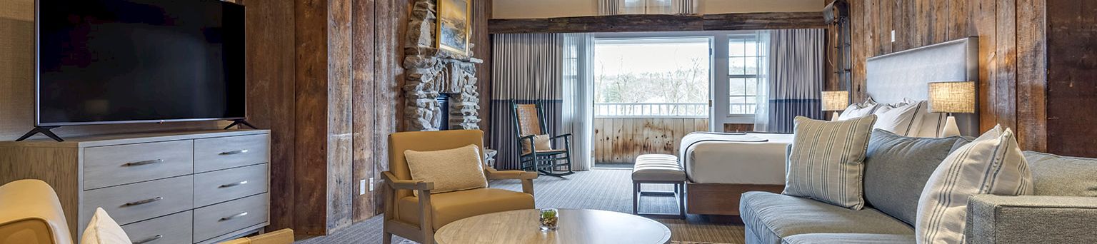 A stylish living room with exposed wooden beam architecture, a blue sofa set, beige chairs, a round coffee table, a TV in a gray cabinet, and a patterned rug.