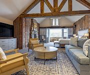 A stylish living room with exposed wooden beam architecture, a blue sofa set, beige chairs, a round coffee table, a TV in a gray cabinet, and a patterned rug.