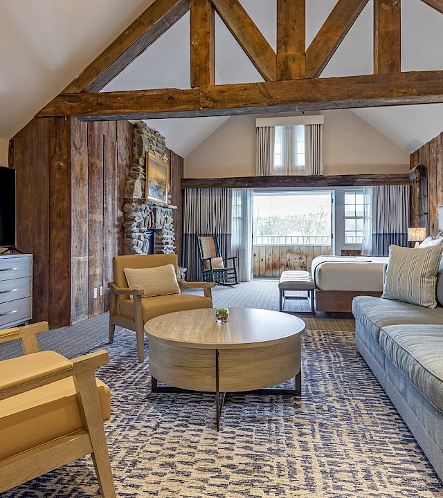 A stylish living room with exposed wooden beam architecture, a blue sofa set, beige chairs, a round coffee table, a TV in a gray cabinet, and a patterned rug.