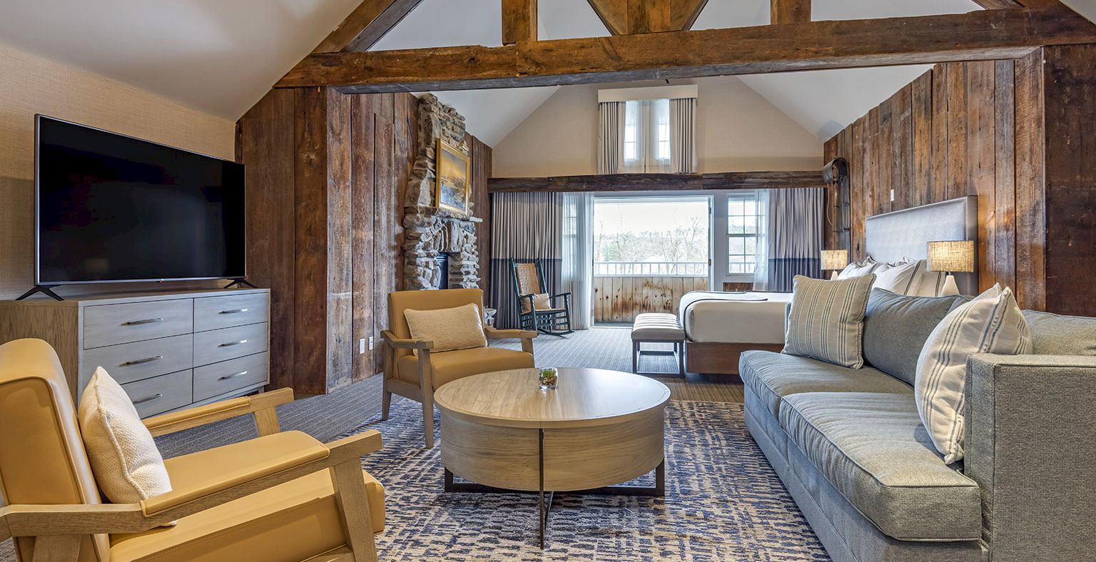 A cozy living room in a rustic loft with exposed wooden beams, a gray sofa, beige chairs, a round coffee table, and a fireplace, bright and inviting.