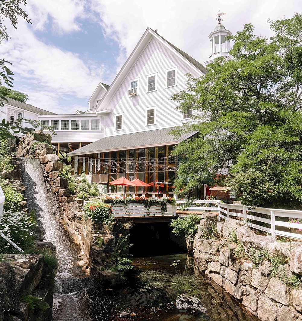 A charming white building perched above a stone-lined stream, with a wooden bridge and outdoor seating under orange umbrellas.